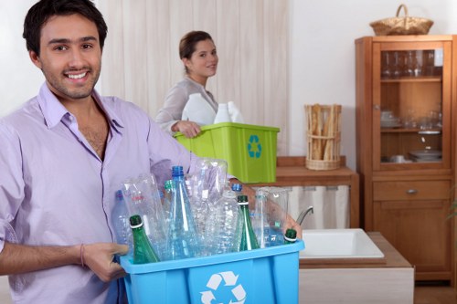 A smiling young man with dark hair and a beard, wearing a light purple shirt, stands in a modern kitchen holding a blue recycling bin filled with empty plastic bottles and containers. Behind him, a woman with dark hair tied back, dressed in a light-colored top, is near a green recycling bin positioned on the kitchen counter. The kitchen features light-colored wooden cabinets, a white tiled backsplash, and a small wooden sideboard with glass doors displaying dishware. To the right, there is a white ceramic sink, and a small area of the kitchen countertop is visible. The environment appears clean, well-lit, and organized, emphasizing household waste management and recycling activities typical of rubbish removal services in Fitzrovia or nearby central London areas, consistent with the services offered by Office Clearance Fitzrovia.