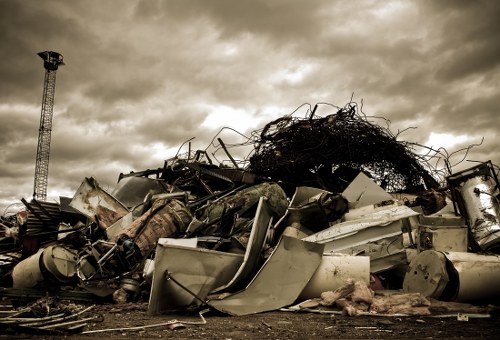A large pile of discarded household and industrial debris is situated outdoors on a concrete or asphalt surface, with various materials including rusted metal, broken appliances, and remnants of furnishings visible. The debris appears to be randomly strewn, with some items leaning against each other and others piled haphazardly. On the upper part of the heap, there is a tangled mass of black and brown wiring or twigs, creating a dense cluster. To the left in the background, a tall metal telecommunications tower with a lattice structure extends upward against a cloudy sky with dark, heavy clouds overhead, suggesting an overcast day. The overall scene is indicative of waste or rubbish removal in an urban environment, possibly in Fitzrovia or nearby areas in London, reflecting a site prepared for clearance by a professional rubbish removal service such as Office Clearance Fitzrovia. The muted colour palette and rough textures highlight the unmanaged state of the debris, emphasizing the need for professional clearance and proper disposal services.