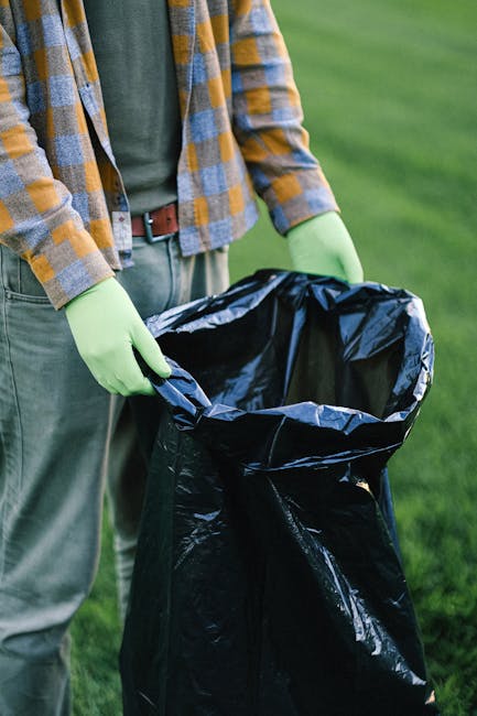 Two waste collection workers are seen beside a large industrial waste truck parked on a city street at dusk. One worker, wearing a dark uniform with reflective orange and yellow safety stripes and the