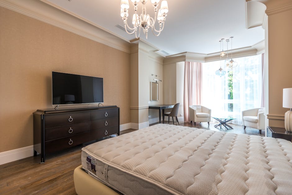 A modern bedroom interior featuring a large, neatly made bed with a white mattress and a beige upholstered headboard positioned against a wall. To the left of the bed, there is a small, dark bedside t