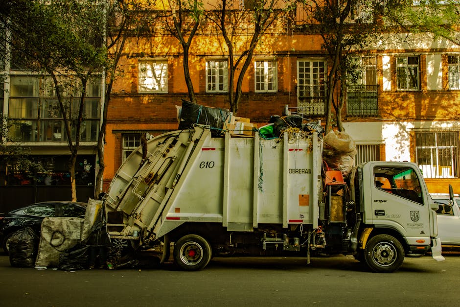 A man with curly dark hair, wearing a black t-shirt and baggy plaid shorts, is standing next to an open, white skip filled with a large pile of discarded roofing tiles, wooden planks, and construction