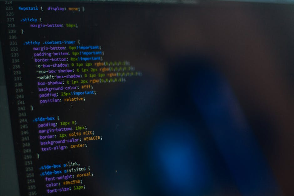 A person in a light blue shirt is seated at a desk, working on a laptop with a black screen displaying colorful lines of code. The individual is holding a black computer mouse with their right hand wh