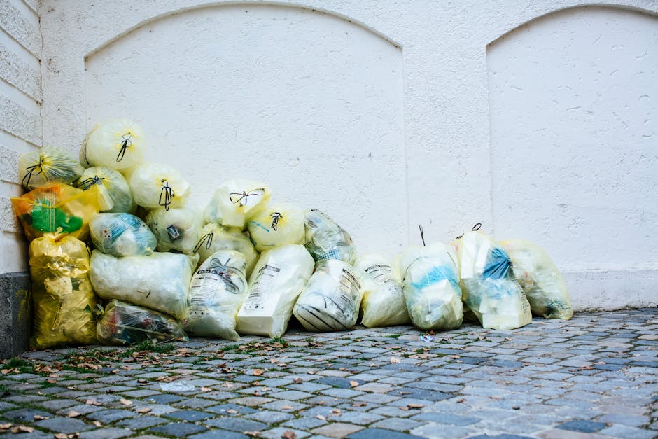 A row of four wheeled waste bins positioned along a cobblestone street in front of a building with an orange facade and three windows fitted with diamond-shaped metal security bars. The first bin on t